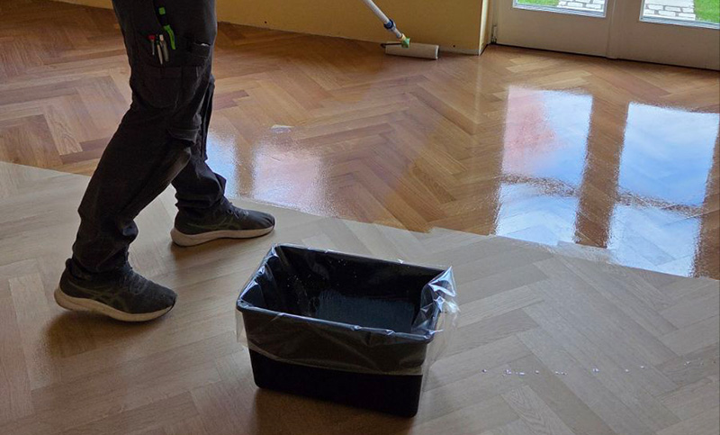 Worker in dark clothing and sneakers rolling a floor roller over a wooden parquet floor; a plastic-lined paint bucket sits nearby.