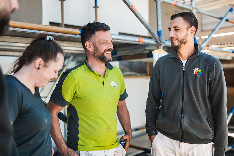 Three construction workers chat and smile near scaffolding on a building site; two men in bright green and dark gray shirts with company logos.