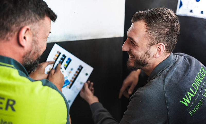 Two coworkers in a workshop compare a color swatch chart, smiling as they discuss it.