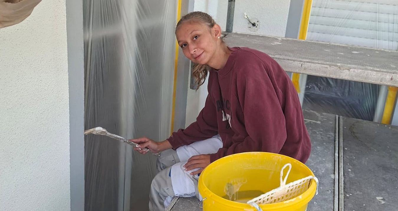 Teenager painting a wall with a small brush, yellow bucket nearby on scaffolding